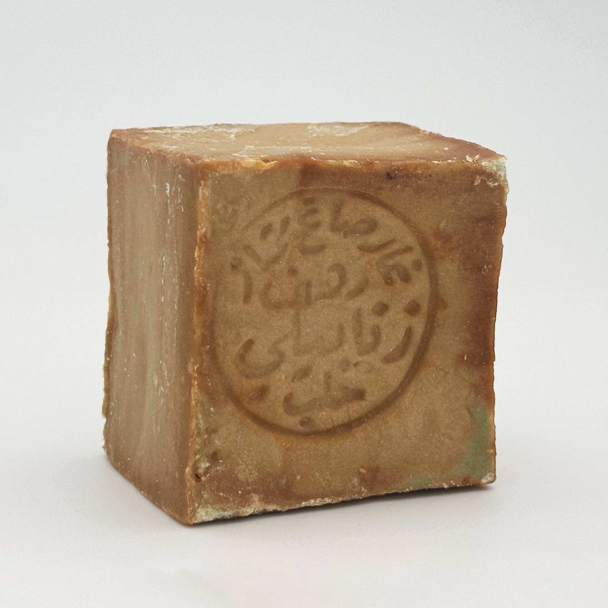 A close-up, angled view of a single square bar of Avlia 100% Olive Oil Aleppo soap, showing its textured, earthy-brown surface and a traditional circular Arabic stamp impressed on the front