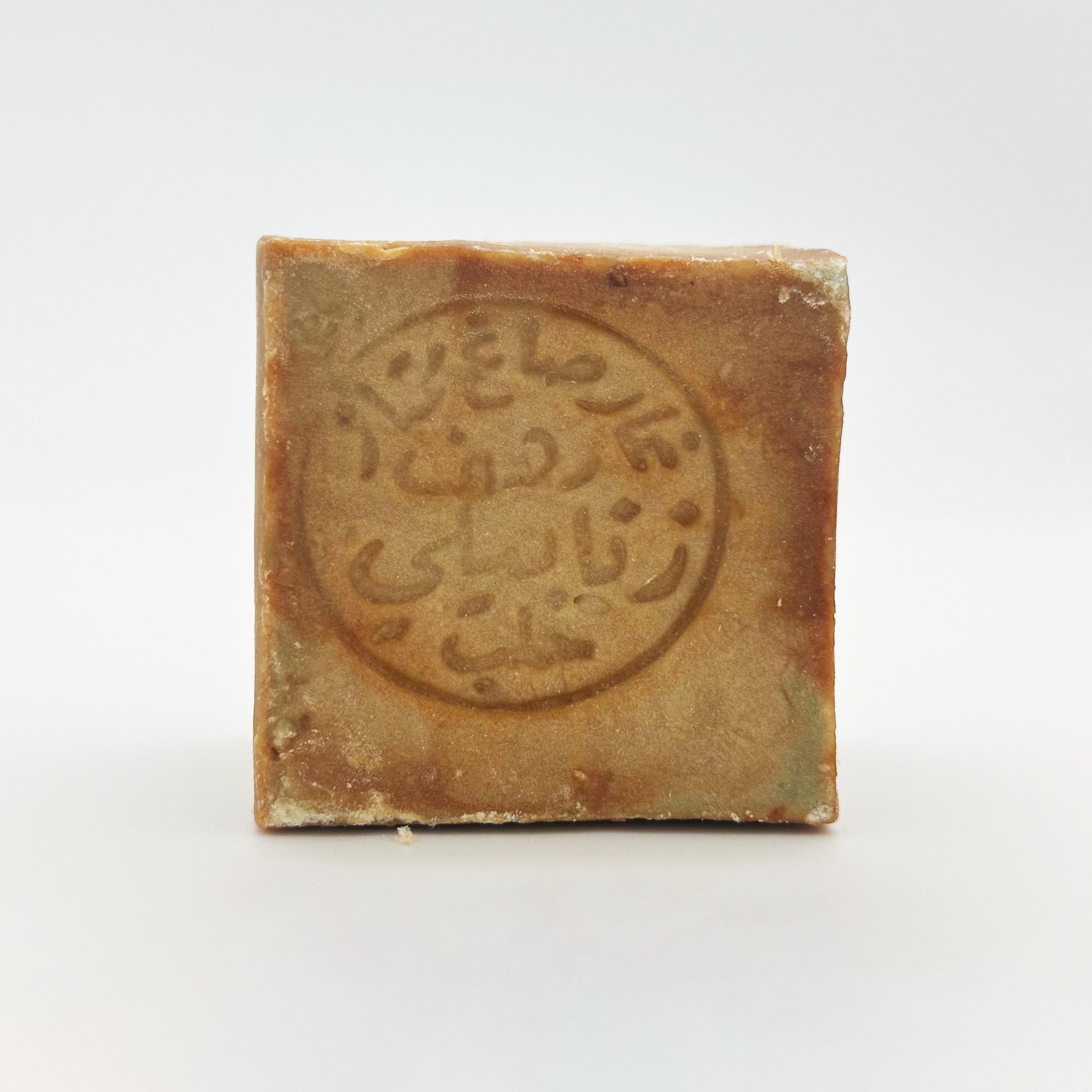 A direct front view of a square Avlia Aleppo soap bar, focused clearly on the detailed circular Arabic calligraphy stamp pressed into the center of the brown surface