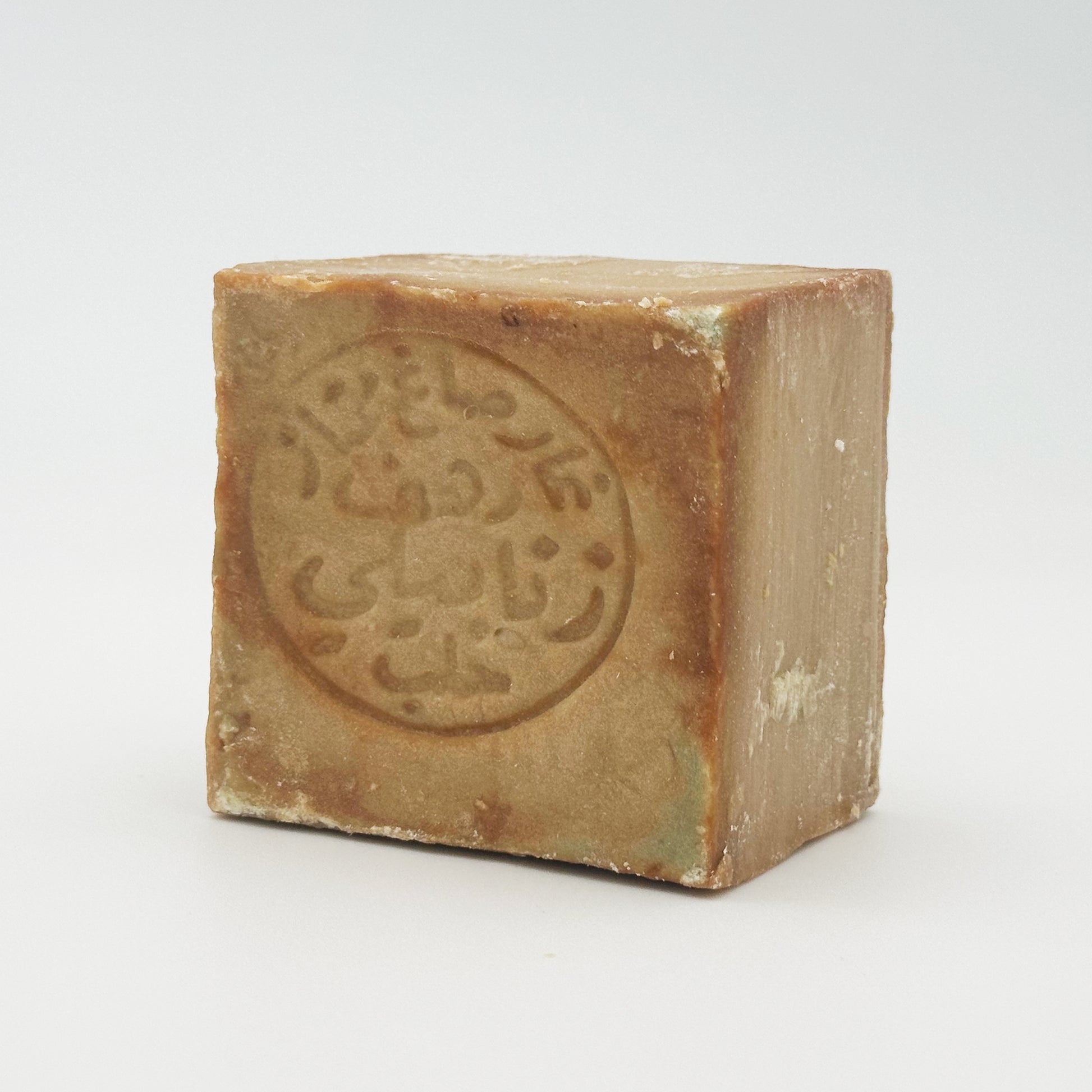 A side-angle view of a thick bar of Avlia 100% Olive Oil Aleppo soap, highlighting the rough, handmade texture of the edges and the stamped design on the front face