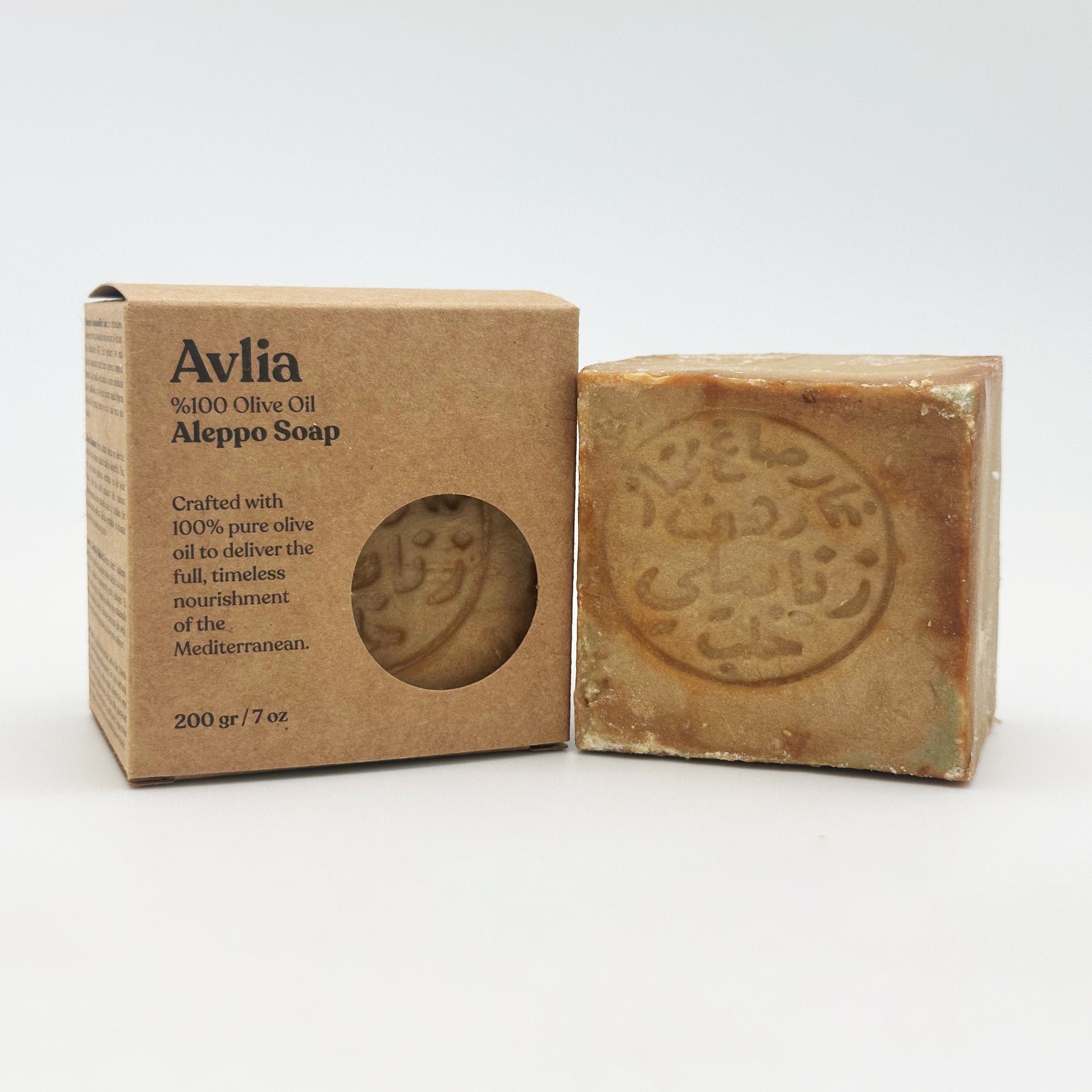 An Avlia brand Aleppo soap bar standing upright next to its brown kraft paper box, which features product text and a circular cutout window revealing the soap inside