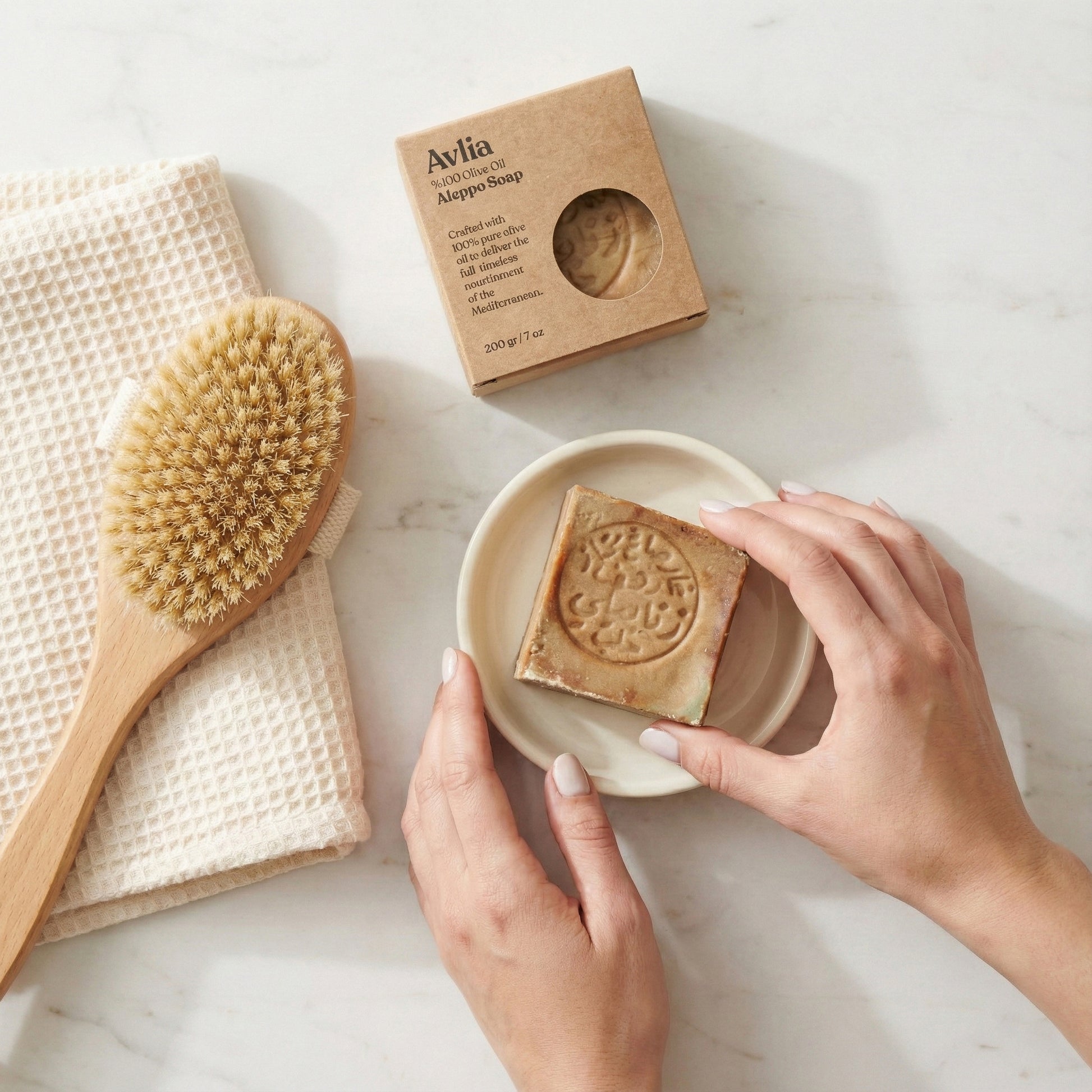 An overhead flat lay photograph shows a pair of hands gently holding a square, stamped Avlia Aleppo soap bar on a small white ceramic dish. To the left, a natural bristle body brush rests on a folded cream waffle towel Portrait