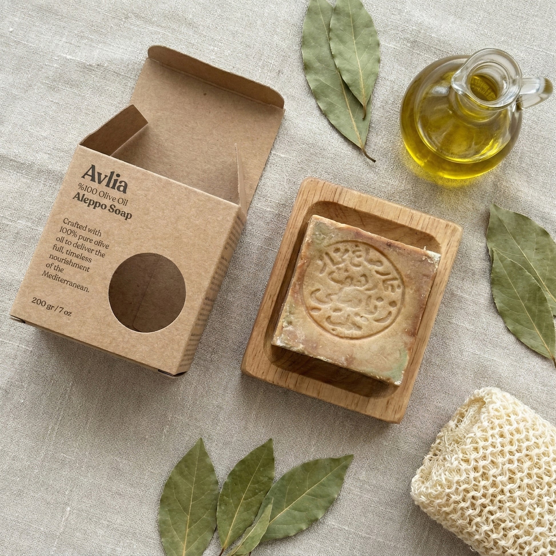 An overhead photograph of natural spa products on a light beige linen surface. In the center, a square, rustic Avlia Aleppo soap bar with an Arabic stamp rests in a wooden dish