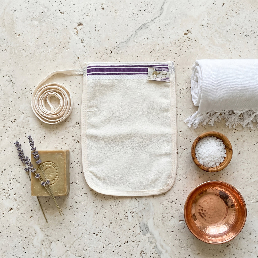 A thin rectangular cream natural Turkish krep kese exfoliating mitt flexible flat pouch shape with softly rounded oval bottom corners on a pale travertine stone surface