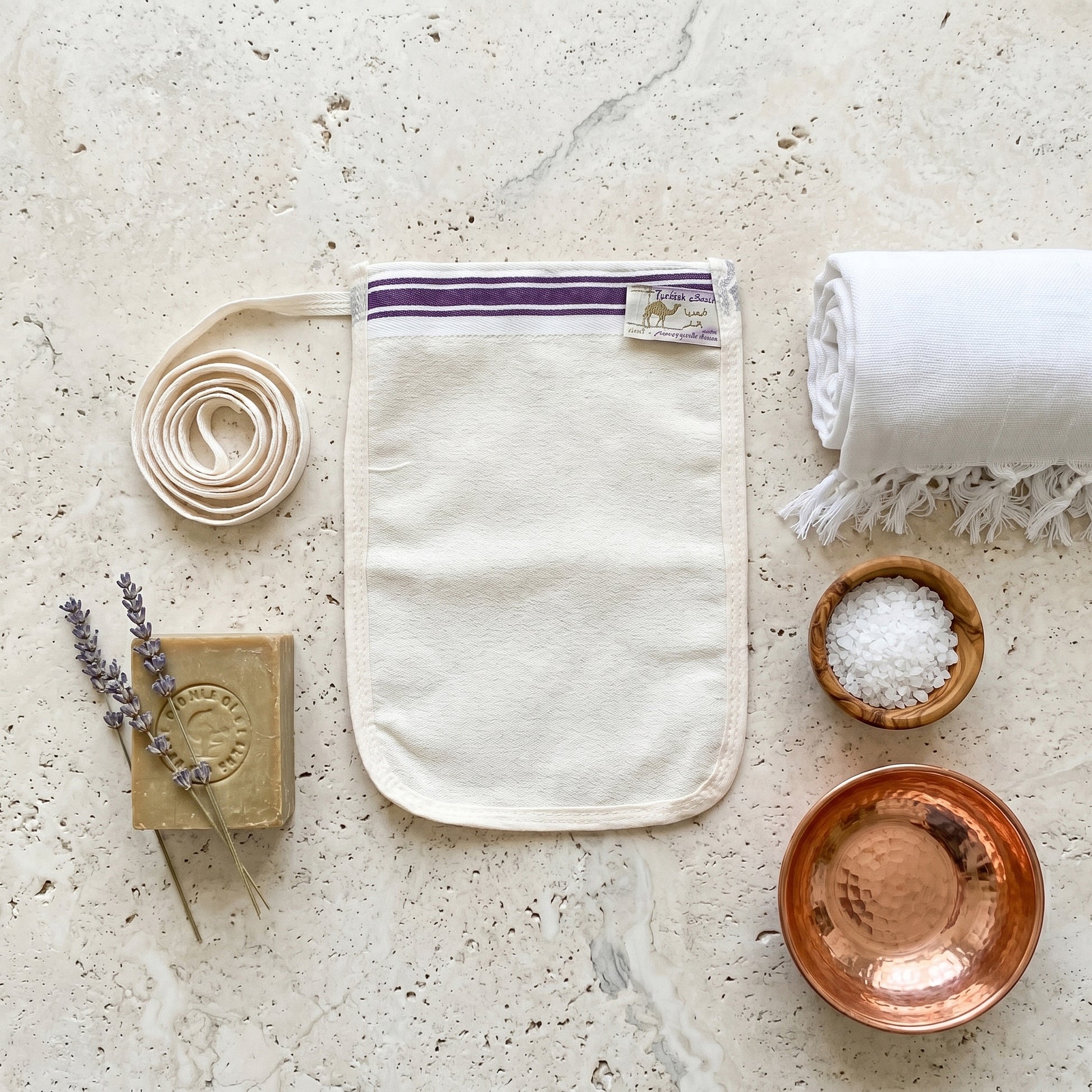 A thin rectangular cream natural Turkish krep kese exfoliating mitt flexible flat pouch shape with softly rounded oval bottom corners on a pale travertine stone surface
