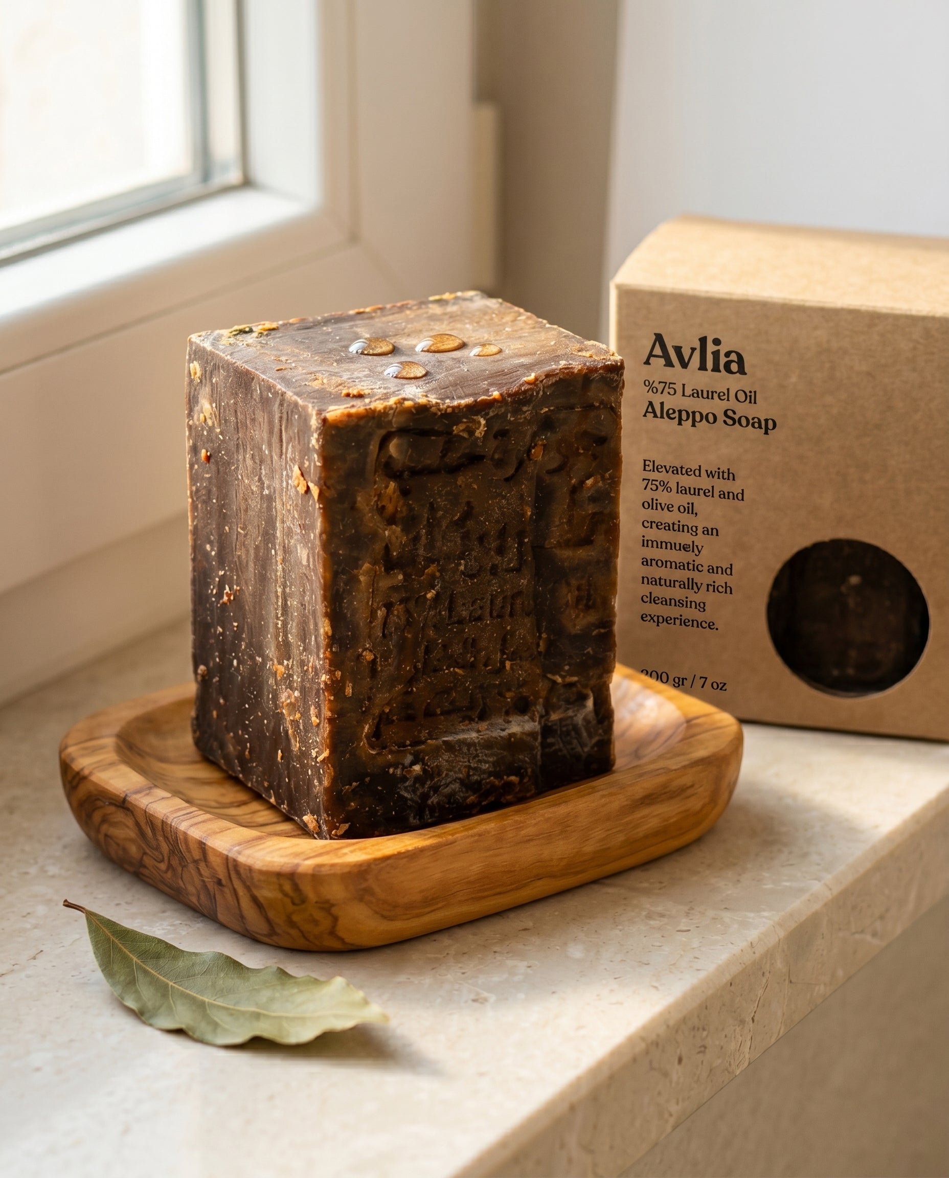 Avlia 75% Laurel Oil Aleppo Soap bar on teak soap dish with bay laurel leaf, dark brown handmade artisan soap, natural morning light bathroom setting
