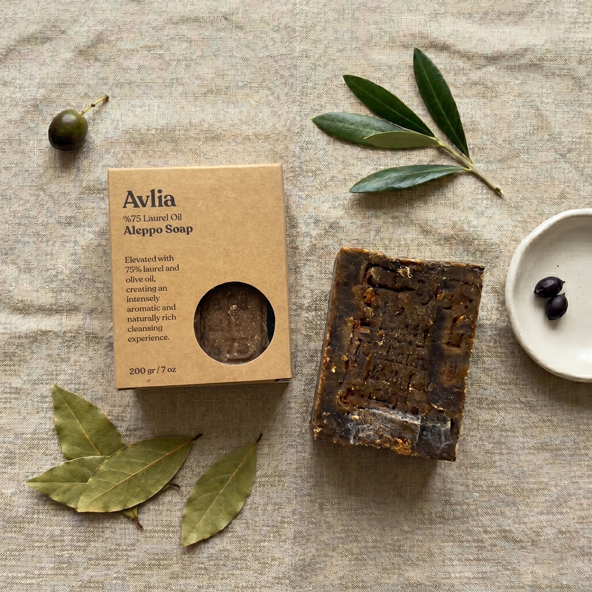 Avlia Aleppo Soap bar and kraft packaging box flat lay overhead with olive branches and bay laurel leaves on linen, natural handmade soap product photography