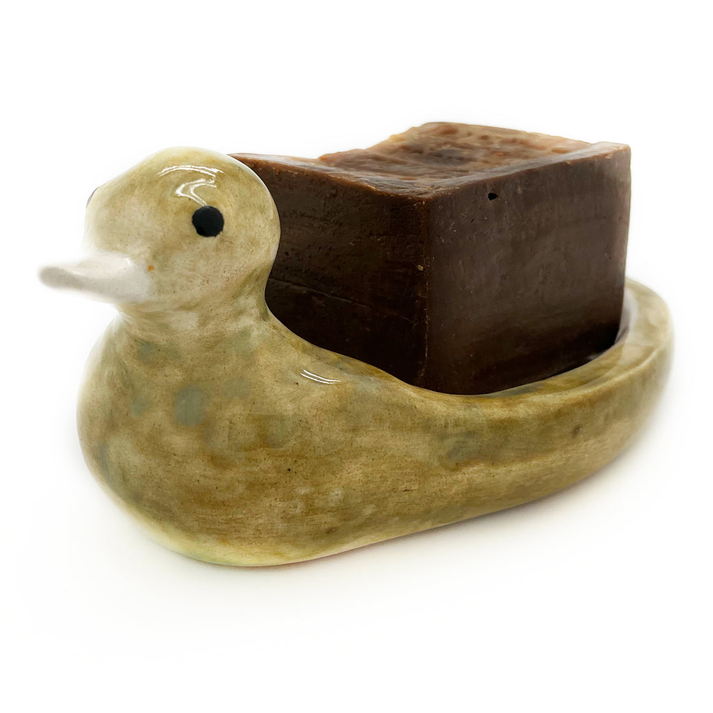 A bar of Aleppo soap resting in the Avlia ceramic duck soap dish, the dark stamped soap contrasting with the cream glazed bowl beneath it