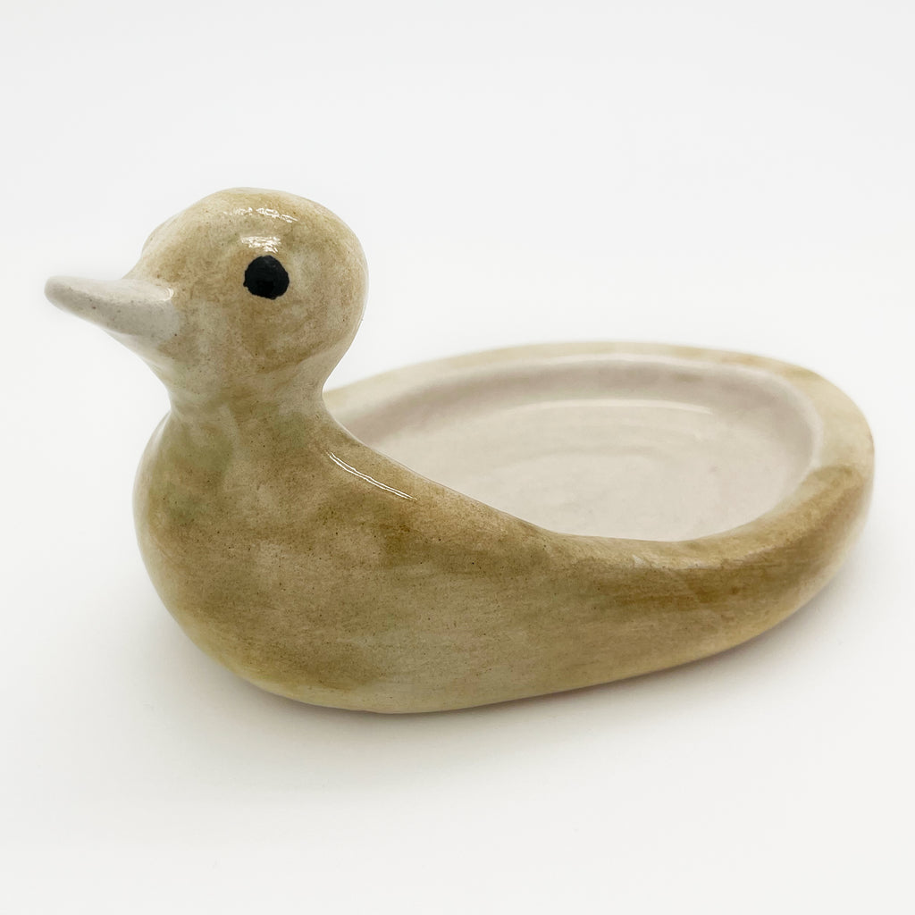 Avlia ceramic duck soap dish in pale cream, three-quarter view with the cream bowl interior and the single black eye dot both visible