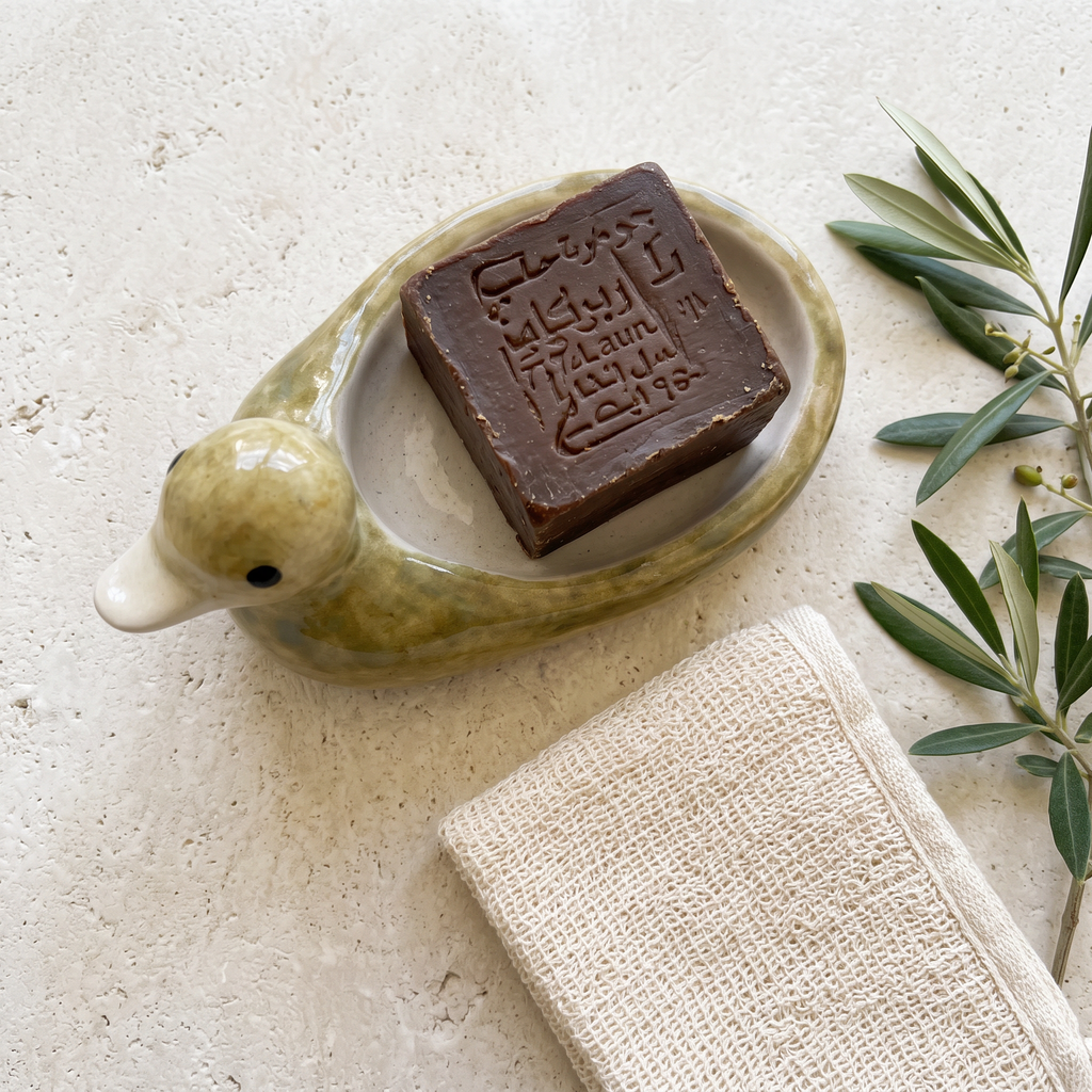 Avlia ceramic duck soap dish photographed from above with Aleppo soap and a folded hammam mitt, arranged on pale limestone with olive leaves