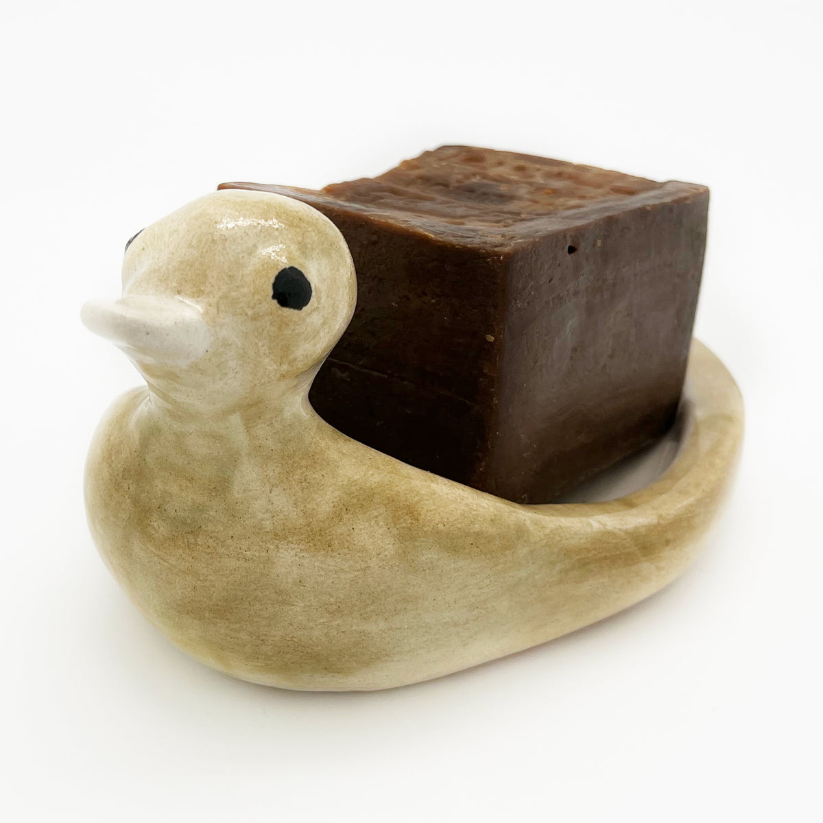 Avlia handmade ceramic duck soap dish in cream glaze holding a stamped bar of Aleppo soap, seen from a low three-quarter angle
