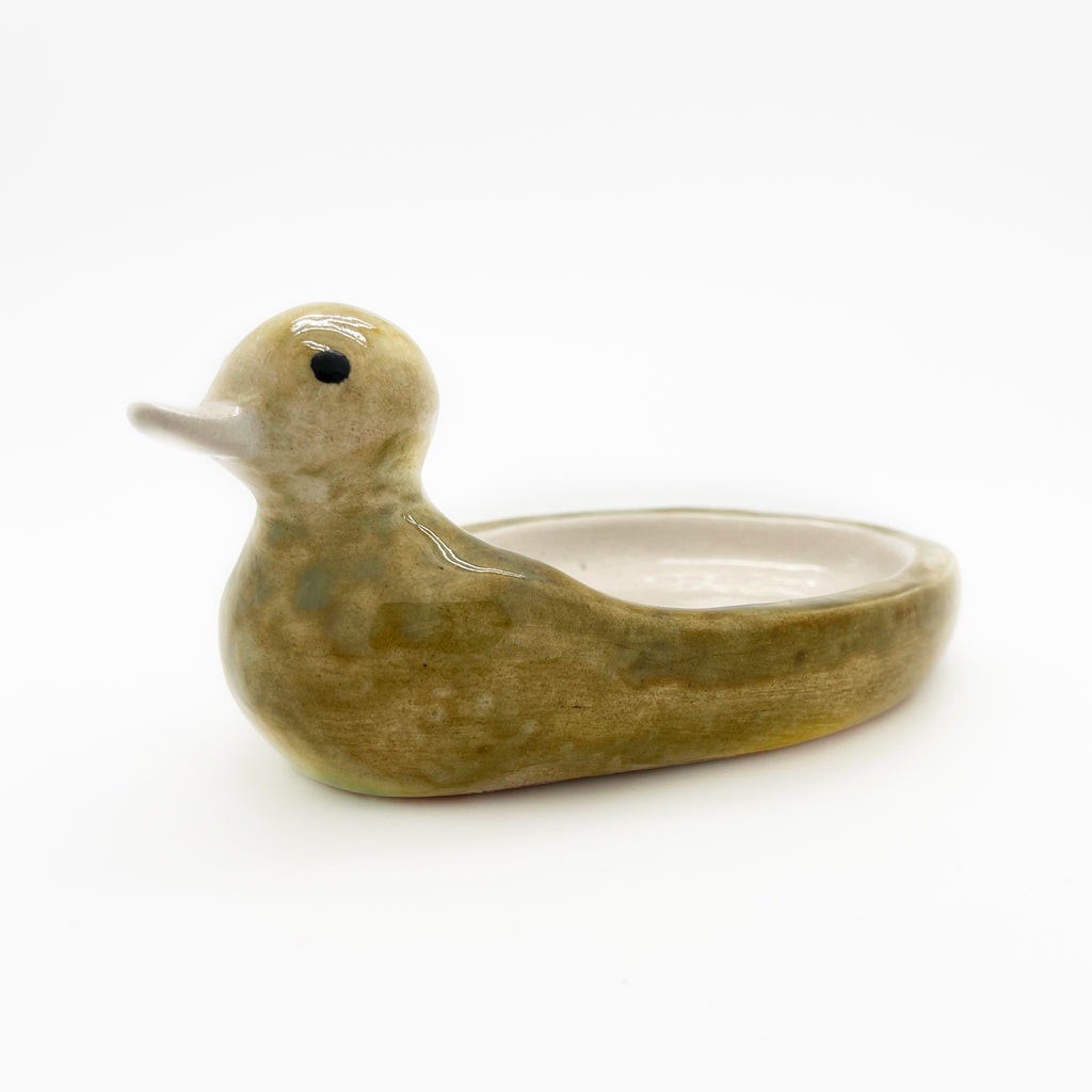 Avlia handmade ceramic duck soap dish in mottled green-mustard glaze, seen from the left at low angle with the cream-glazed bowl hollowed into its back