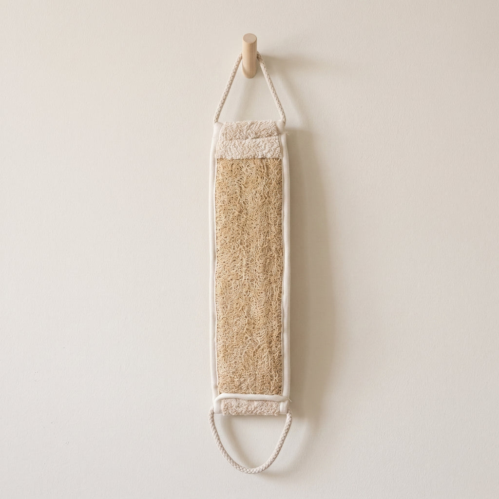 Avlia natural luffa back strap hanging by one braided cotton loop from a hook on a pale wall, tan luffa face and cream terry band both visible
