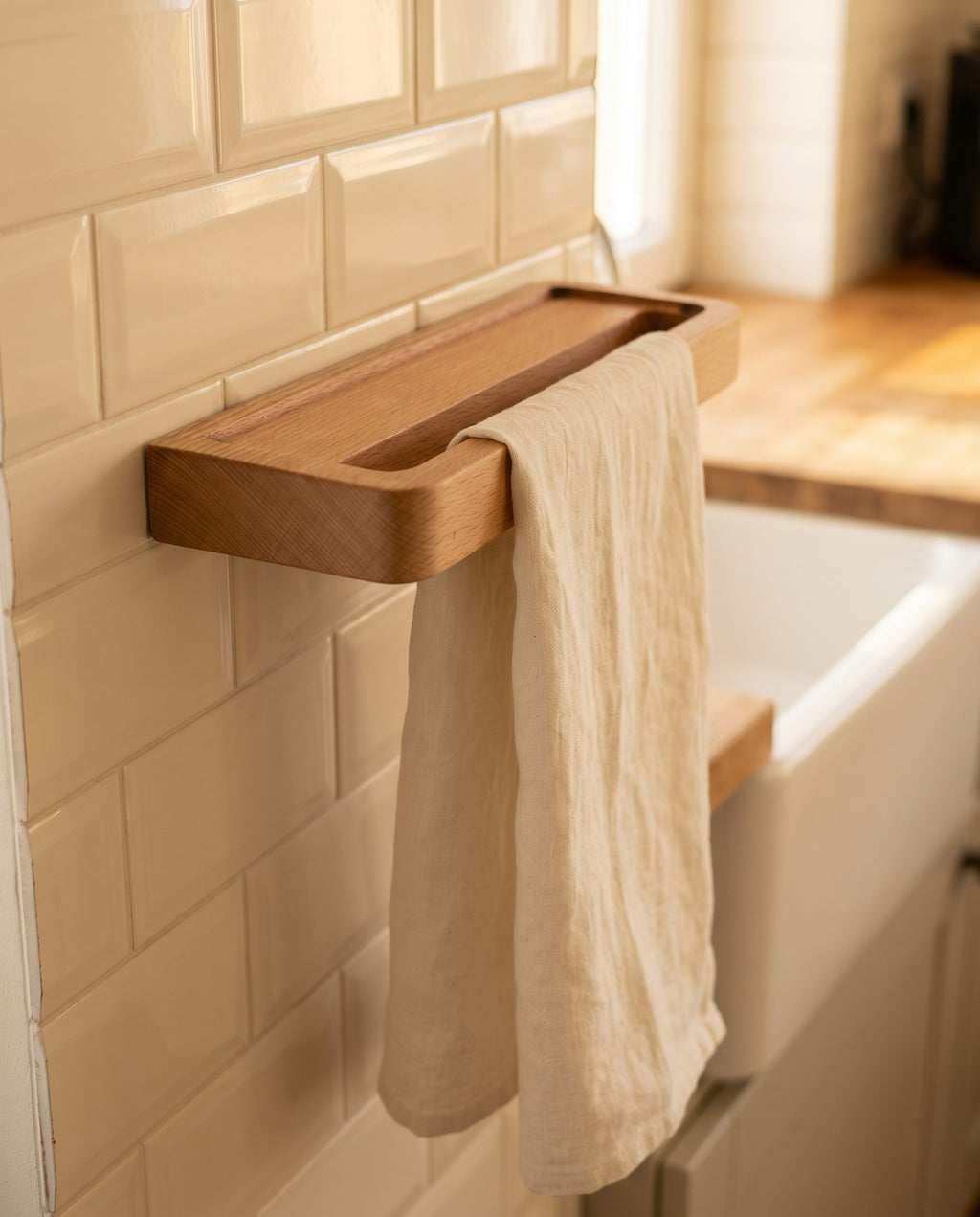 Avlia beechwood towel shelf mounted on kitchen wall beside farmhouse sink, cream linen tea towel hanging below through slot, floating wooden kitchen accessory