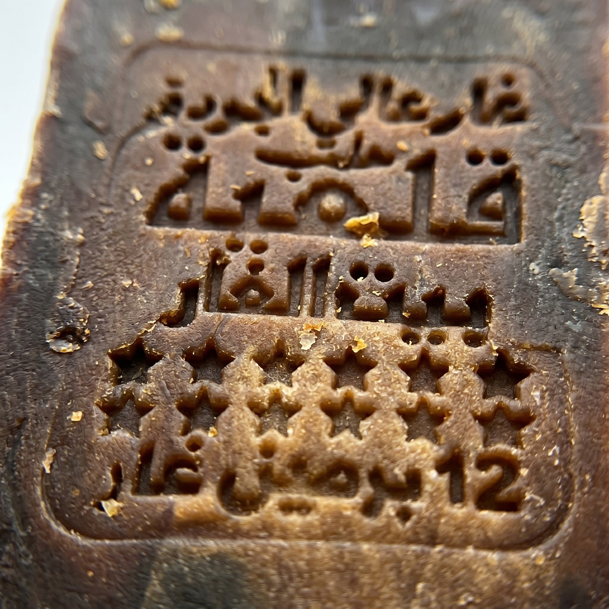 Extreme close-up of traditional Arabic stamp impression on Avlia 50% Laurel Oil Aleppo Soap surface, centuries-old hand-pressed production seal on handmade dark brown bar