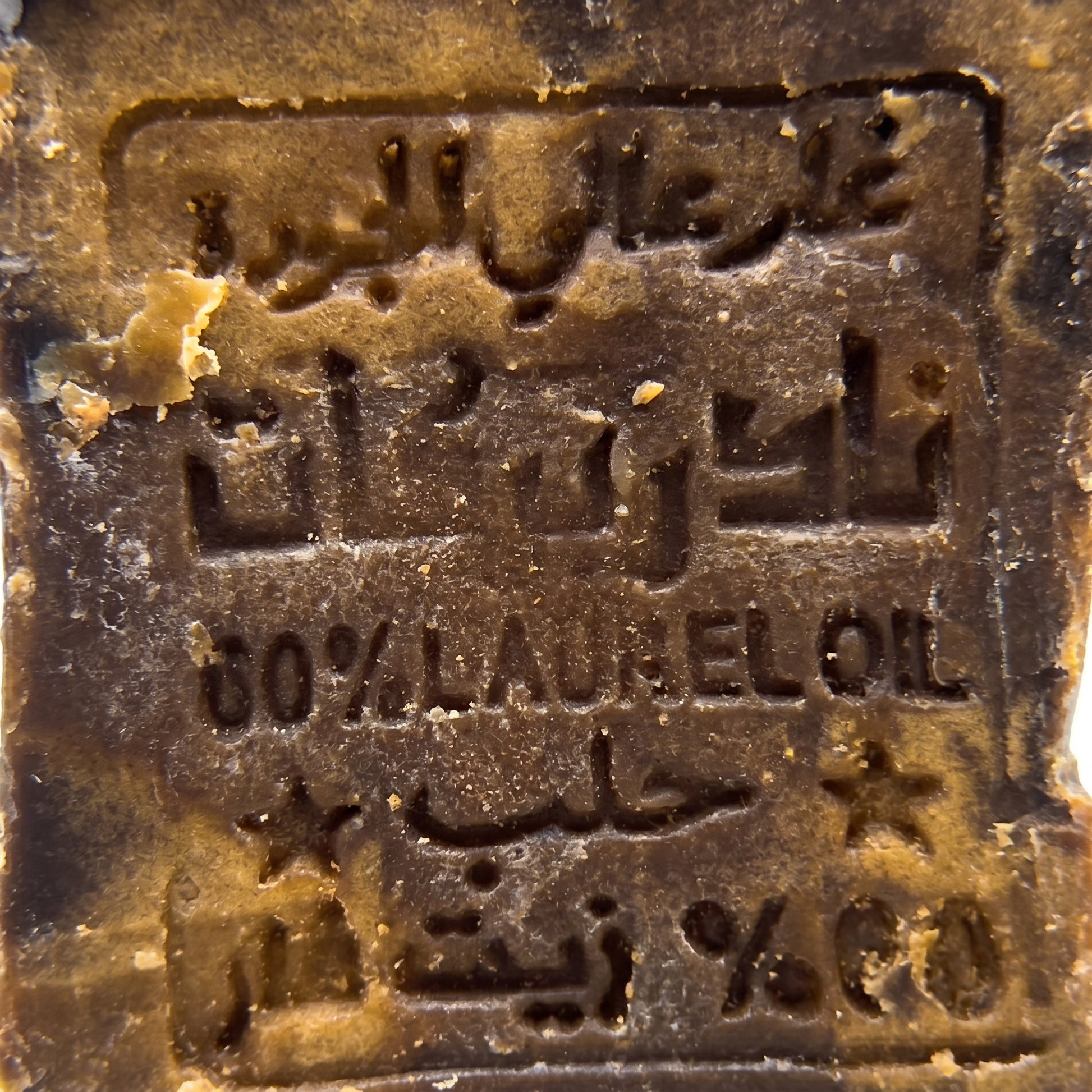 Extreme close-up of traditional Arabic stamp impression on Avlia 60% Laurel Oil Aleppo Soap surface, centuries-old hand-pressed production seal on handmade dark brown bar