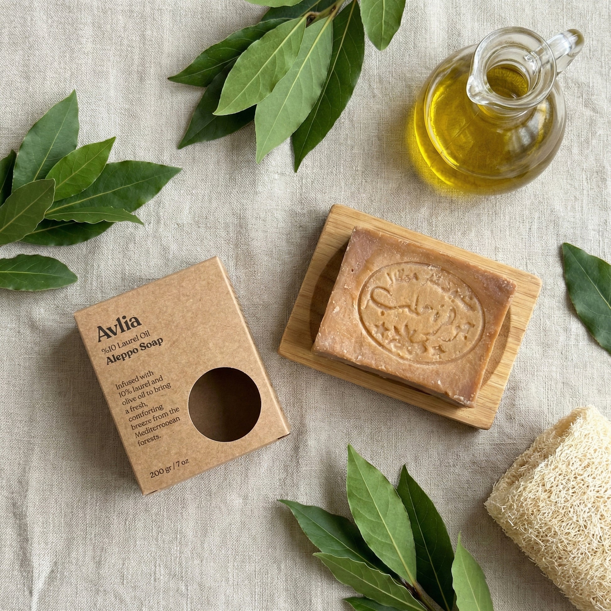 Flat lay composition showing Avlia 10% Laurel Oil Aleppo Soap bar on a wooden dish, its kraft packaging box, a glass cruet of olive oil, fresh laurel leaves, and a natural loofah sponge on a beige linen background