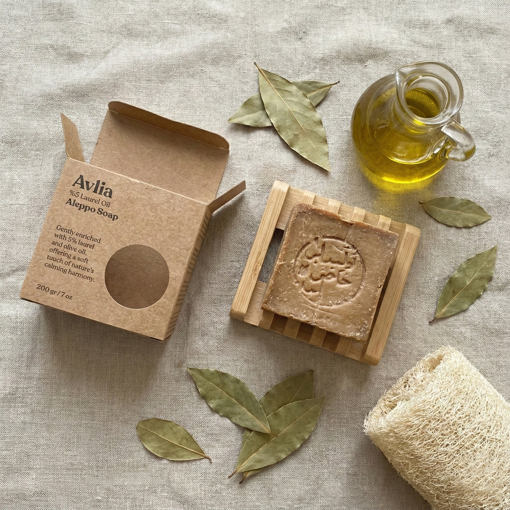 Flat lay of Avlia Aleppo soap bar on a wooden dish, open cardboard packaging, a cruet of olive oil, bay leaves, and a loofah on a linen surface