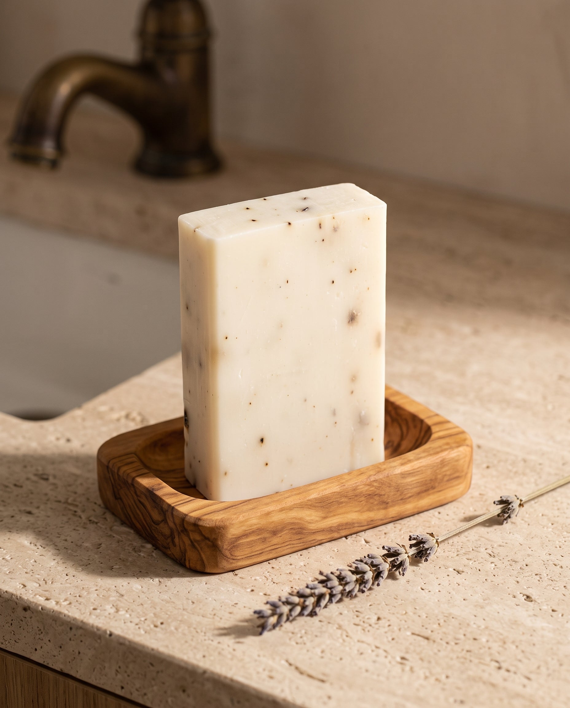 Lavender Soap Bar in an olive wood soap dish beside a dried lavender stem on a stone vanity