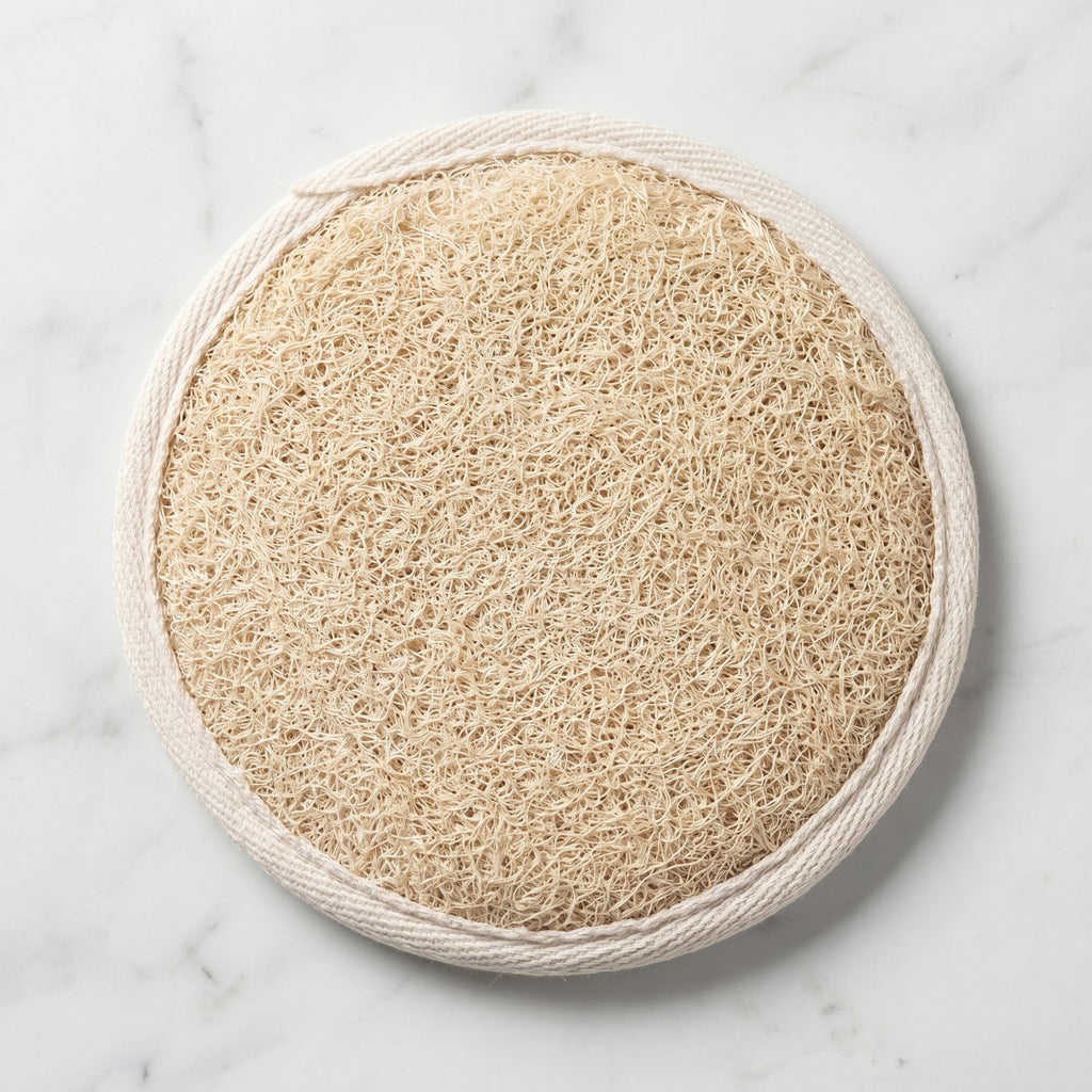 Natural loofah pad on white marble, round Luffa aegyptiaca exfoliating sponge with cream cotton trim, hero shot