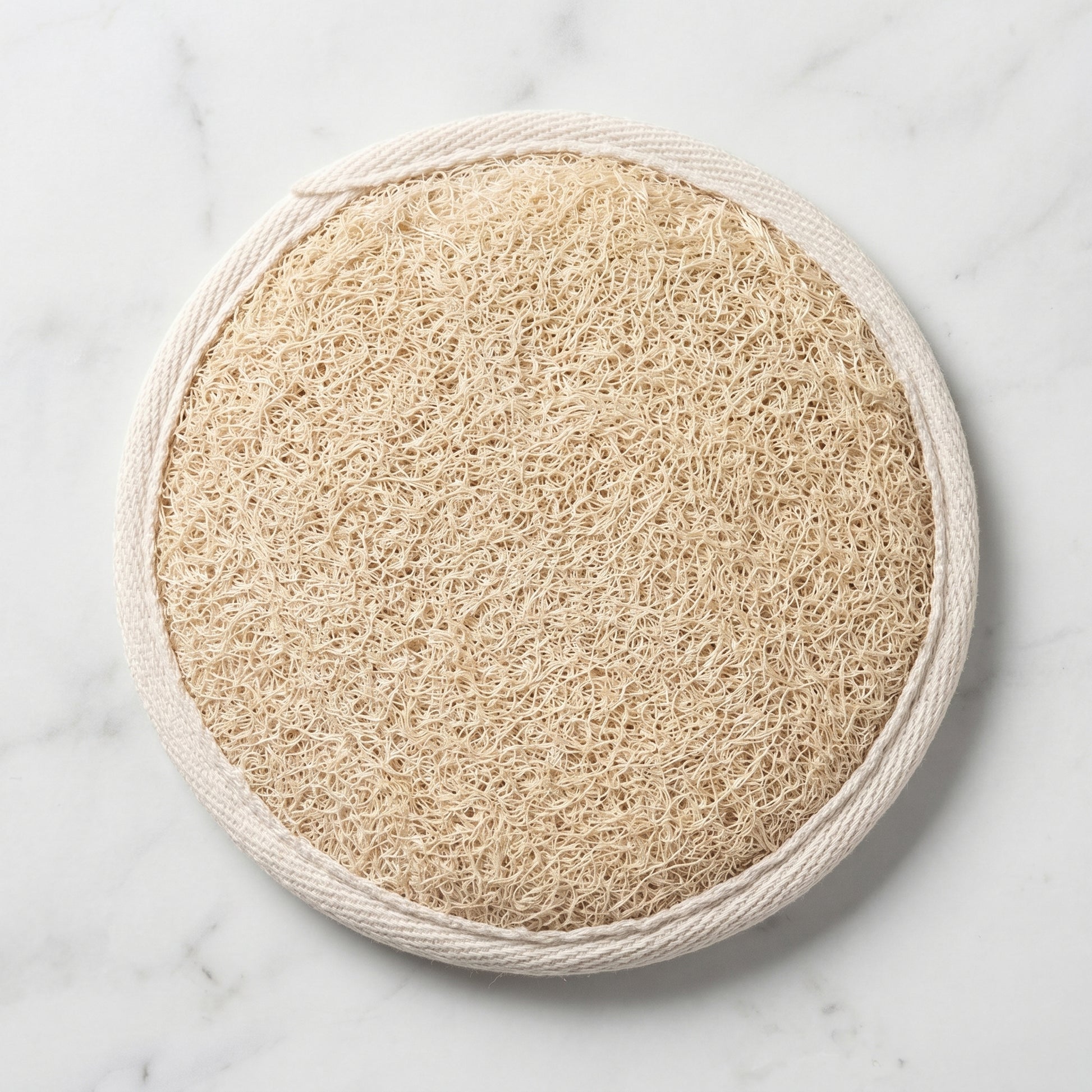 Natural loofah pad on white marble, round Luffa aegyptiaca exfoliating sponge with cream cotton trim, hero shot