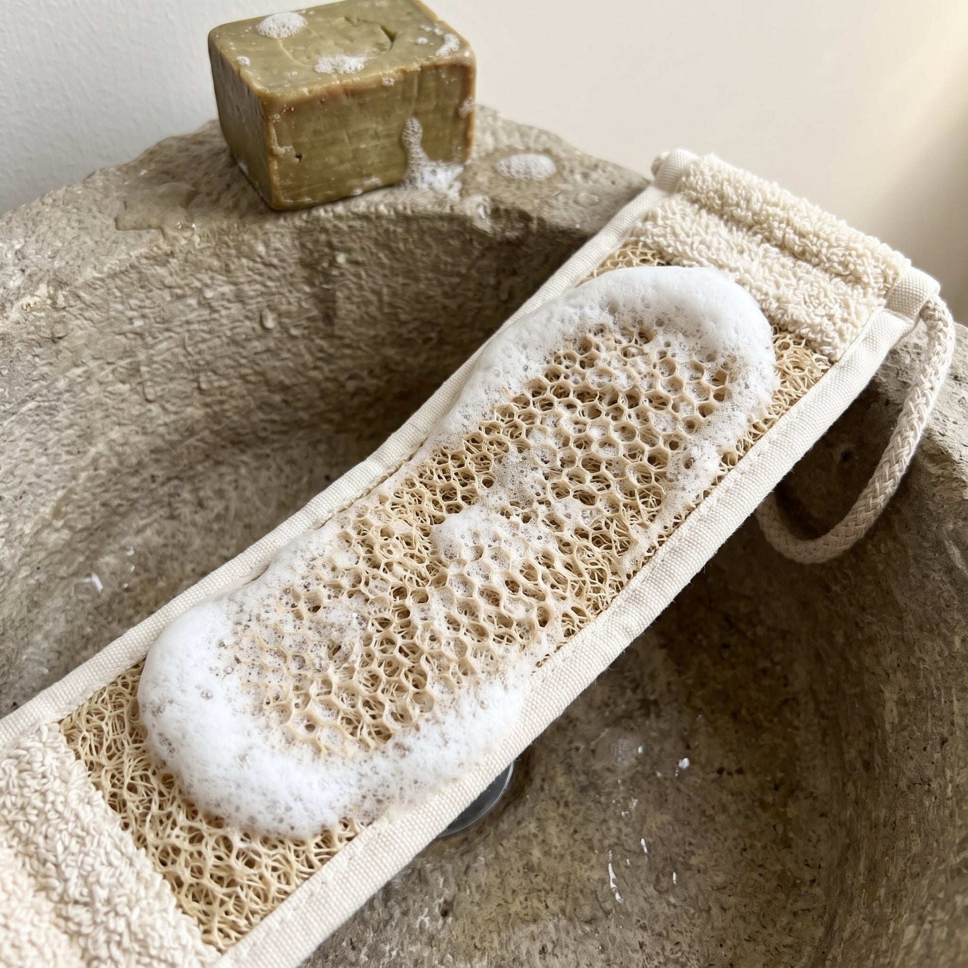 The luffa face of the Avlia back strap worked into thick white lather over a stone basin, beside a part-used olive oil soap bar