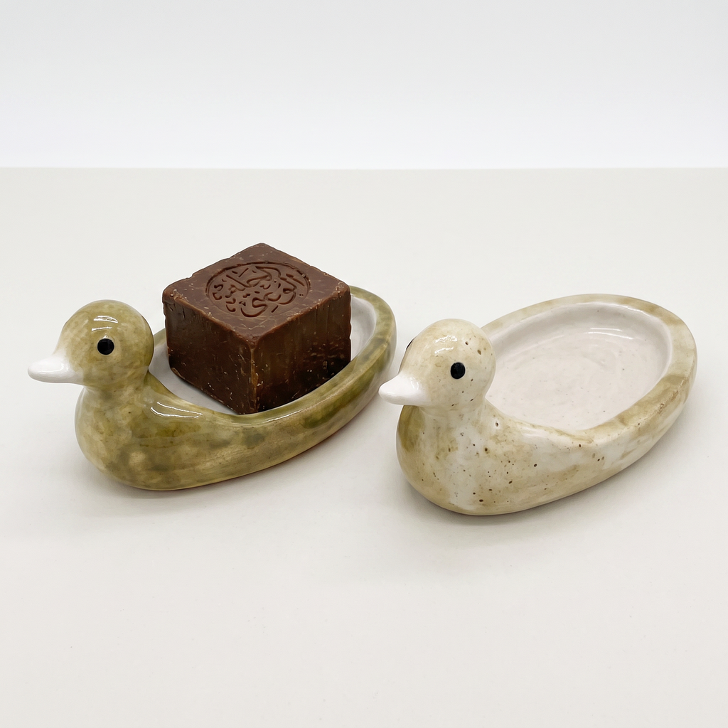 Two Avlia ceramic duck soap dishes side by side, one in green-mustard glaze and one in pale cream, showing how the glaze varies between pieces