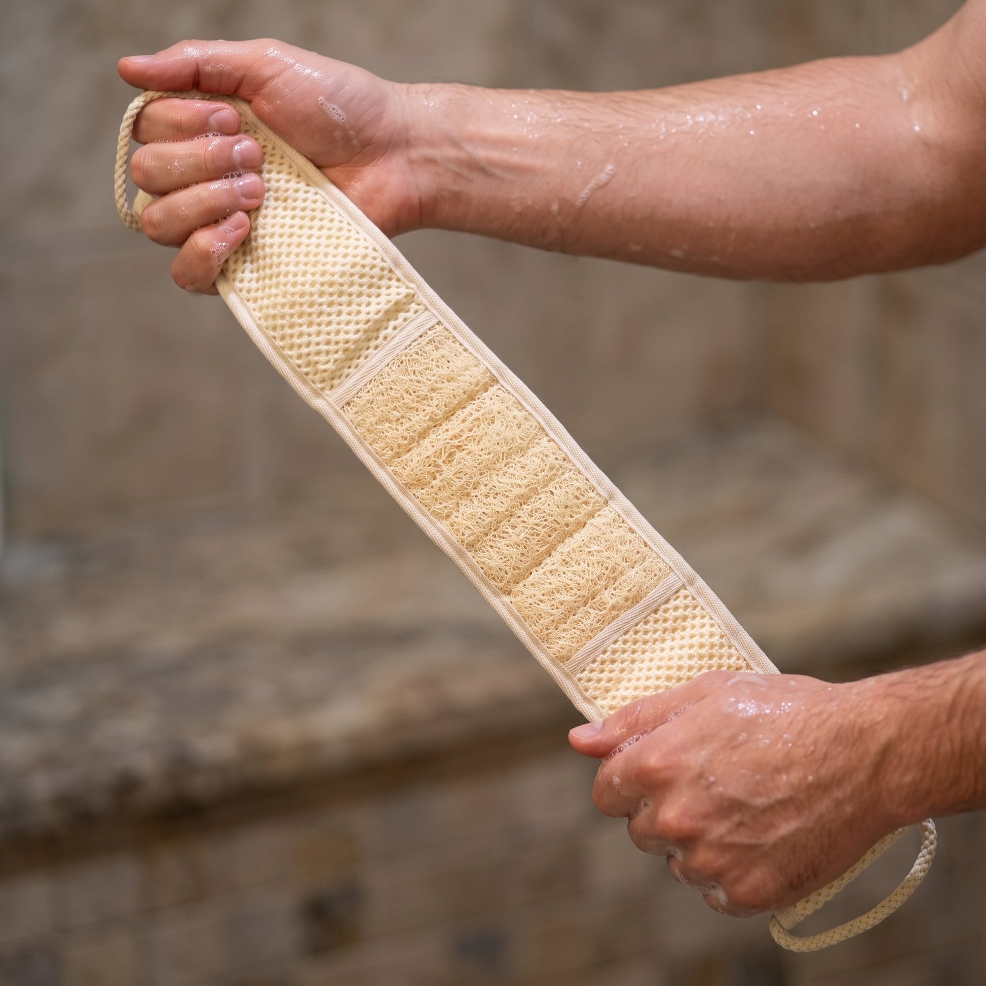 Two hands gripping the braided cotton cord loops of the Avlia back strap, the strap held taut and diagonal between them, wet skin and lather