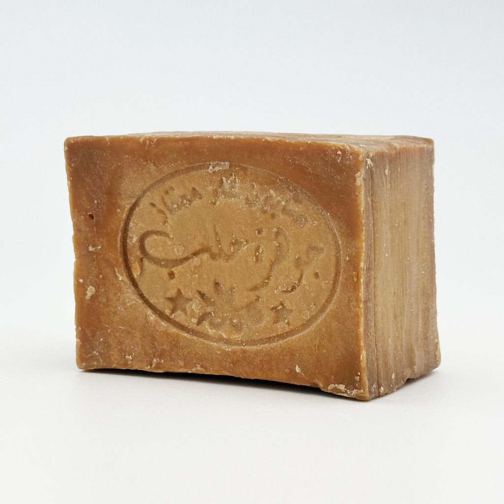 Unpackaged bar of Avlia Aleppo soap showing rustic texture and traditional Arabic stamp impression, front angled view Portrait