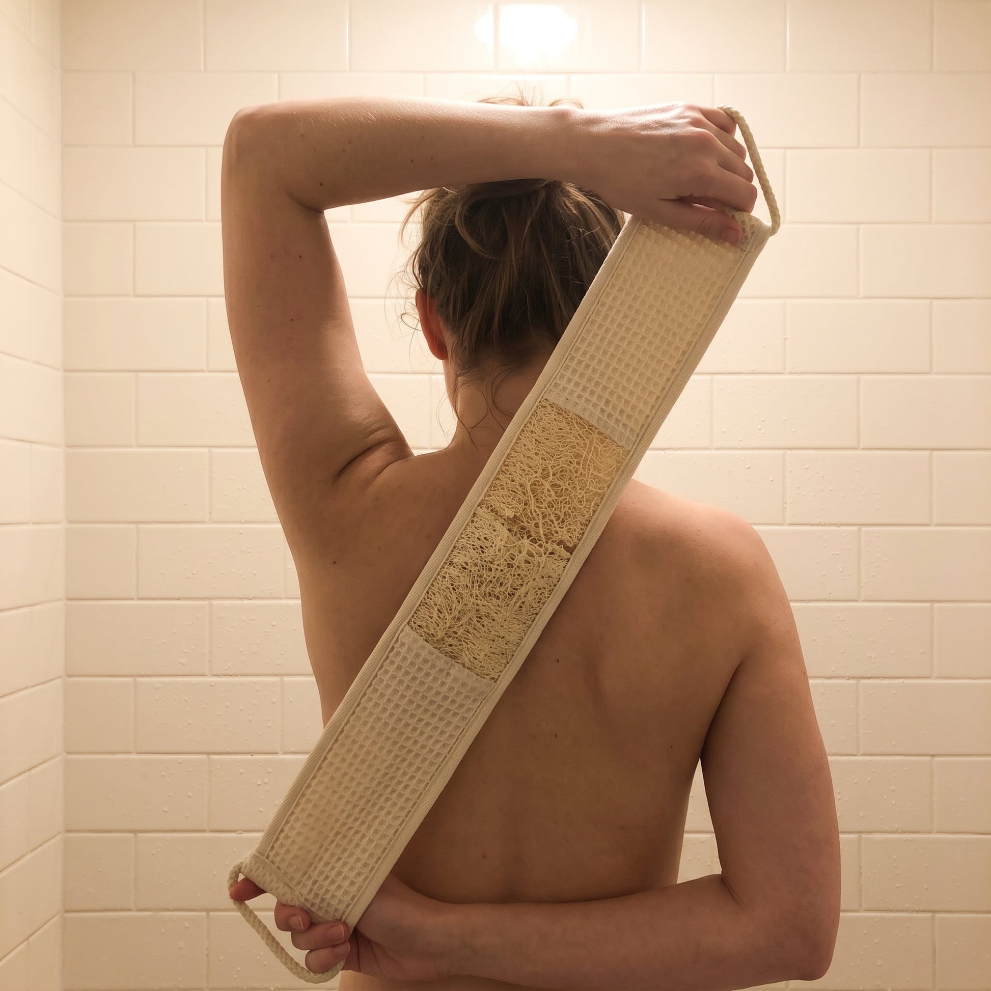Woman pulling the Avlia extra long loofah back strap diagonally across her upper back, one hand raised behind her neck and one low at her waist