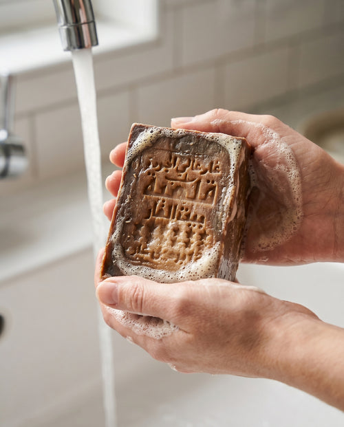 Woman's hands lathering Avlia 50% Laurel Oil Aleppo Soap bar under running water, creamy natural lather from olive oil and laurel berry oil handmade soap